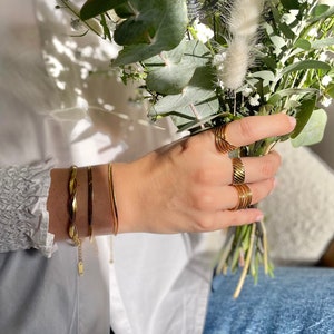 K&ouml;nnte beinhalten: Eine Hand tr&auml;gt einen Satz goldener Ringe und zwei goldene Armb&auml;nder. Die Hand h&auml;lt einen Strau&szlig; gr&uuml;ner und wei&szlig;er Blumen.