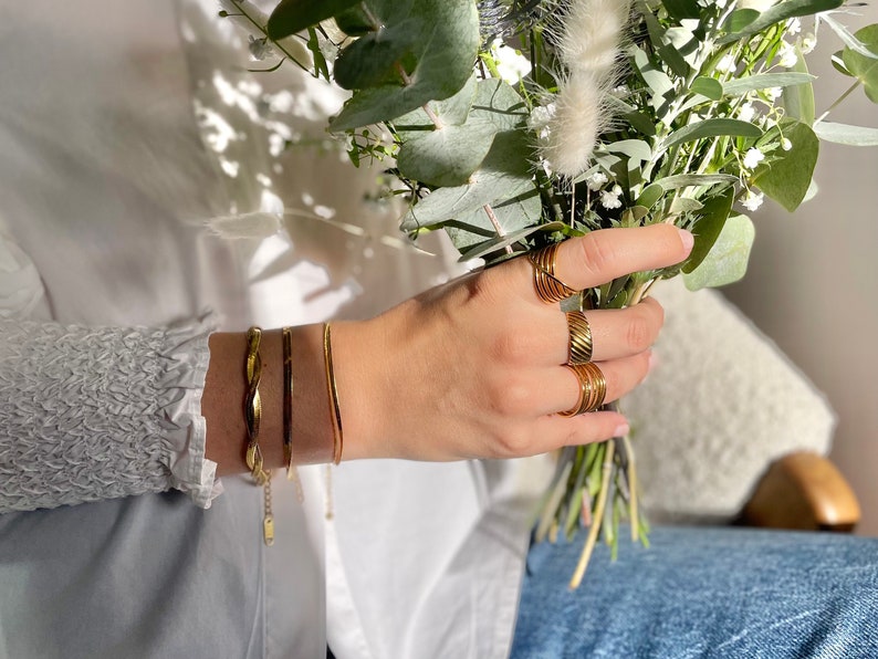 K&ouml;nnte beinhalten: Eine Hand tr&auml;gt einen Satz goldener Ringe und zwei goldene Armb&auml;nder. Die Hand h&auml;lt einen Strau&szlig; gr&uuml;ner und wei&szlig;er Blumen.