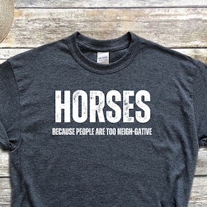 May include: A dark gray t-shirt with the text "Horses because people are too neigh-gative" in white lettering. The shirt is laid out on a wooden surface with a pair of black sneakers, a straw hat, a pair of denim shorts, a black watch, and white earbuds.