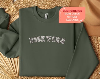 Pode incluir: Uma camisola verde escura com o texto "BOOKWORM" bordado em branco na frente. A camisola está colocada sobre uma superfície tecida.