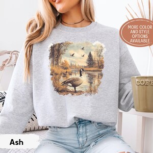 Retro fall scene with a goose by the lake and geese flying overhead, printed on a cozy 90s-style nature crewneck sweatshirt or unisex t-shirt, perfect for bird lovers and wildlife gift giving