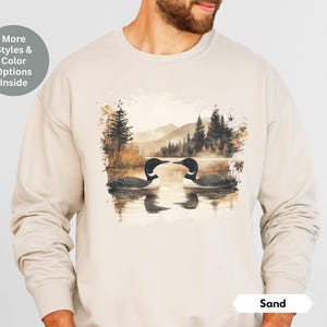 Cozy sand-colored sweatshirt featuring a pair of loons on a serene lake with a retro watercolor-style forest and mountain background. Perfect for nature lovers or as a thoughtful vintage-inspired gift.