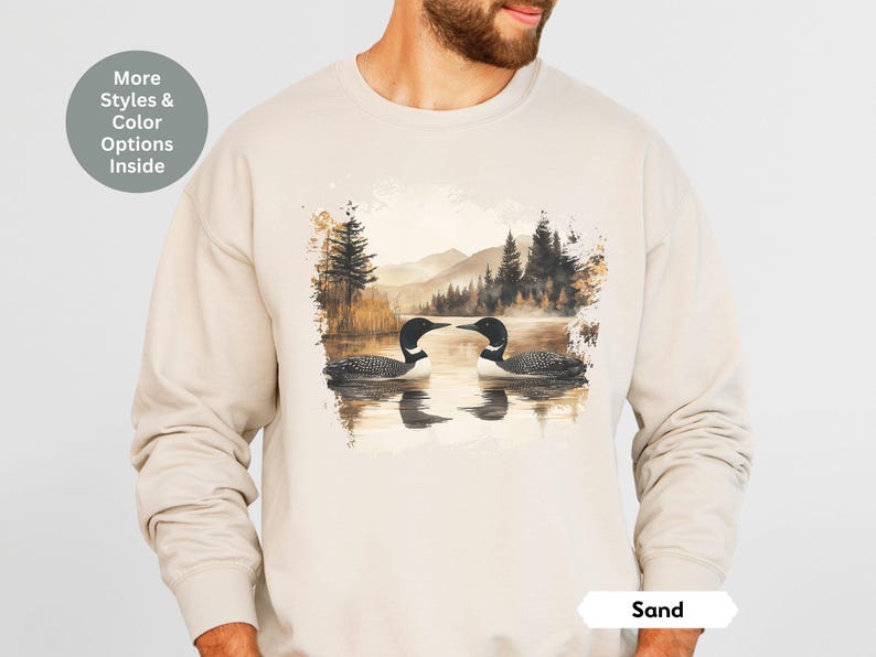 Cozy sand-colored sweatshirt featuring a pair of loons on a serene lake with a retro watercolor-style forest and mountain background. Perfect for nature lovers or as a thoughtful vintage-inspired gift.