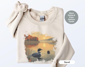 May include: Sand-colored sweatshirt featuring a scenic lake design with a loon, reeds, and a sunset. The graphic includes a loon swimming in the water, with the text "More Styles & Color Options Inside" and "Sand".
