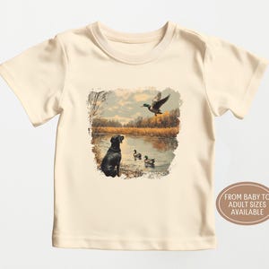 Retro duck hunting t-shirt featuring a black lab and mallard ducks by a pond, printed outdoors scene for babies, kids, and adults, perfect for hunting families and dog lovers.