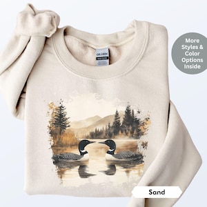 Cozy sand-colored sweatshirt featuring a pair of loons on a serene lake with a retro watercolor-style forest and mountain background. Perfect for nature lovers or as a thoughtful vintage-inspired gift.