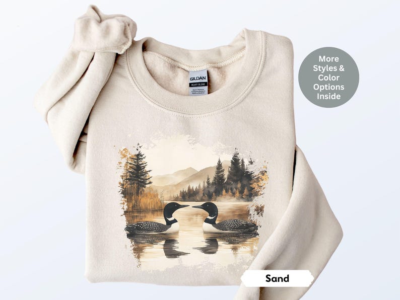 Cozy sand-colored sweatshirt featuring a pair of loons on a serene lake with a retro watercolor-style forest and mountain background. Perfect for nature lovers or as a thoughtful vintage-inspired gift.