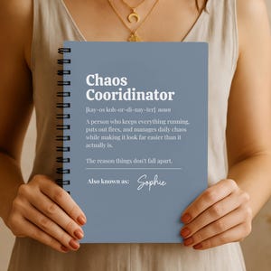 May include: A spiral-bound notebook with a blue cover. The cover features the words "Chaos Coordinator" in white, along with a definition and the name "Sophie." The notebook is held by a person.