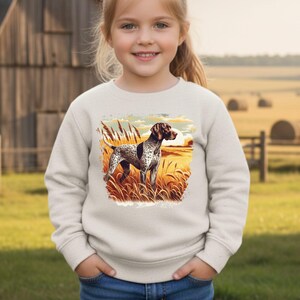 May include: A cream-colored sweatshirt featuring a detailed illustration of a German Shorthaired Pointer dog standing in a field of tall grass. The dog has a brown and white spotted coat. The background shows a sunset over a field.