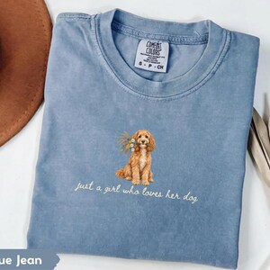May include: A blue jean colored t-shirt with a crew neckline and a Comfort Colors brand tag. The center features an embroidered golden-brown dog holding a bouquet of colorful flowers, with the text "just a girl who loves her dog" below.