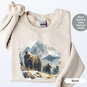 May include: A beige sweatshirt with a graphic print of two brown bears in a snowy mountain landscape. The text "Sand" is visible in the lower right corner of the image.