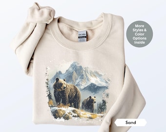 Può includere: Una felpa beige con una stampa grafica di due orsi bruni in un paesaggio montano innevato. Il testo "Sand" è visibile nell'angolo in basso a destra dell'immagine.