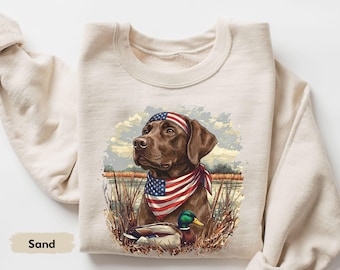 Può includere: Felpa color sabbia con un cane marrone che indossa una bandana e una fascia per la testa rosse, bianche e blu. La grafica include un'anatra e una scena paesaggistica. La parola "Sand" è stampata in basso a sinistra.
