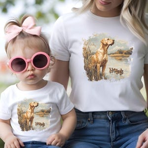 Retro duck hunting t-shirt featuring a yellow lab standing by a pond watching mallard ducks, printed design available in baby, toddler, youth, and adult sizes