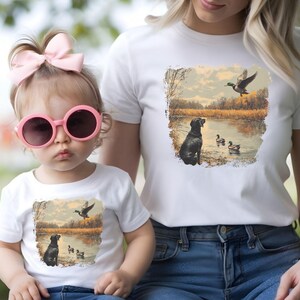 Retro duck hunting tshirt featuring a black lab and mallard ducks by a pond, printed outdoors scene for babies, kids, and adults, perfect for hunting families and dog lovers.
