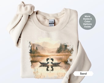 May include: A beige sweatshirt with a watercolor design of two loons kissing in a lake with a mountain range in the background. The text "Sand" is printed on the bottom right corner of the sweatshirt.