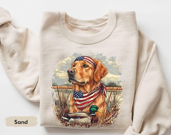 Può includere: Felpa color sabbia con una grafica di un Golden Retriever con bandana e fascia con la bandiera americana. Un'anatra è in primo piano. Il design è ambientato sullo sfondo di un paesaggio paludoso.