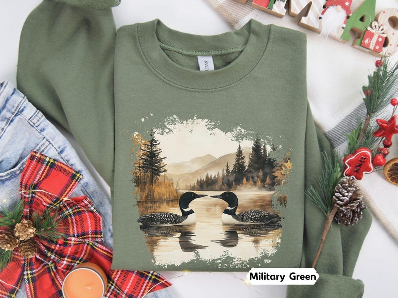Cozy military green colored sweatshirt featuring a pair of loons on a serene lake with a retro watercolor-style forest and mountain background. Perfect for nature lovers or as a thoughtful vintage-inspired gift.