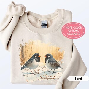 May include: A beige sweatshirt with a watercolor illustration of two birds in a snowy forest. The text "More Color Options Available" is in a pink circle on the right side of the sweatshirt. The text "Sand" is in a white rectangle on the bottom right side of the sweatshirt.