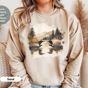 Cozy sand-colored sweatshirt featuring a pair of loons on a serene lake with a retro watercolor-style forest and mountain background. Perfect for nature lovers or as a thoughtful vintage-inspired gift.
