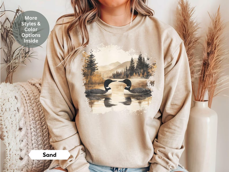 Cozy sand-colored sweatshirt featuring a pair of loons on a serene lake with a retro watercolor-style forest and mountain background. Perfect for nature lovers or as a thoughtful vintage-inspired gift.