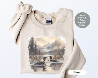 May include: A sand-colored sweatshirt with a wintery scene of a lake, mountains, and two loons. The text "Sand" is visible in the bottom right corner.