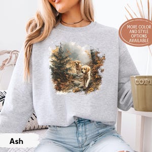May include: Light gray sweatshirt featuring a watercolor print of two golden retrievers walking on a forest path. The sweatshirt has the word "Ash" printed on the bottom. The text "MORE COLOR AND STYLE OPTIONS AVAILABLE" is also visible.