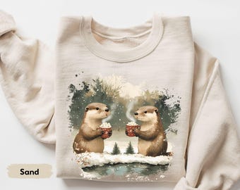 May include: Sand-colored sweatshirt featuring an illustration of two otters holding mugs of hot chocolate in a snowy winter scene. The otters are brown with white accents, and the mugs are red with white designs.