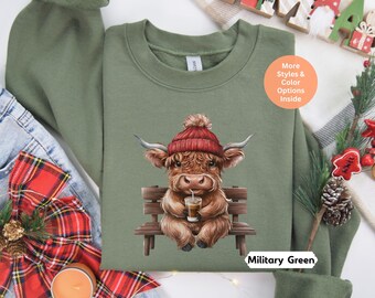 May include: A green sweatshirt with a cartoon illustration of a Highland cow wearing a red Santa hat and holding a cup of coffee. The cow is sitting on a wooden bench. The sweatshirt has the text "Military Green" printed on the bottom right corner.