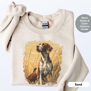 May include: A beige sweatshirt with a brown and white dog and a pheasant in a field printed on the front. The text "Sand" is printed on the bottom right corner of the image.