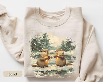 May include: Sand-colored sweatshirt featuring an illustration of two otters holding a string of lights in a snowy winter scene. The otters are brown with light-colored bellies, and the background includes snow-covered trees and a stream.