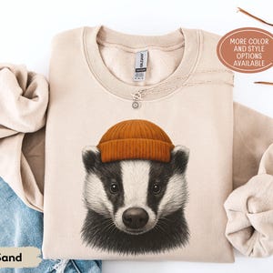 May include: Sand-colored sweatshirt featuring a badger wearing an orange beanie. The badger has black and white fur and a detailed illustration. The sweatshirt has a crew neck and long sleeves. The text "MORE COLOR AND STYLE OPTIONS AVAILABLE" is in a brown oval.