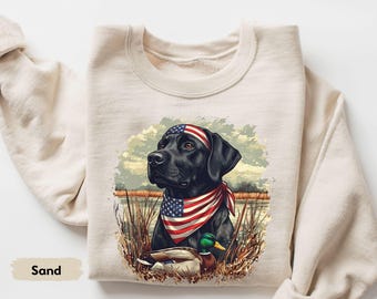Può includere: Felpa color sabbia con una grafica di un Labrador nero che indossa una bandana e una fascia con un disegno della bandiera americana. Un'anatra è in primo piano. La felpa è uno stile casual e confortevole.