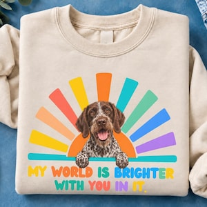 Peut inclure: Un sweat-shirt beige à col rond orné d'un motif représentant un chien tacheté marron et blanc, les pattes posées sur une ligne horizontale. Derrière le chien se trouve un soleil levant multicolore. Le texte indique, My world is brighter with you in it.
