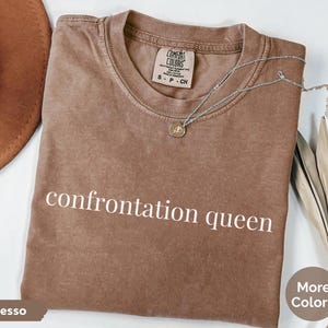 May include: Espresso-colored t-shirt with the text "confrontation queen" in white. A silver necklace with a pendant rests on the shirt. The shirt is folded, with a brown hat and dried plant in the background.