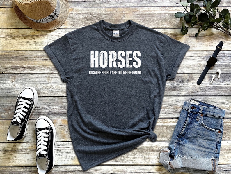 May include: A dark gray t-shirt with the text "Horses because people are too neigh-gative" in white lettering. The shirt is laid out on a wooden surface with a pair of black sneakers, a straw hat, a pair of denim shorts, a black watch, and white earbuds.