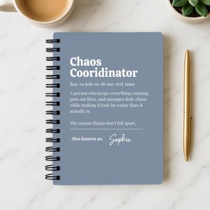 May include: A spiral-bound notebook with a blue cover, featuring the text "Chaos Coordinator" and its definition. A gold pen and a cup of coffee are next to the notebook. A small plant is also in the image.