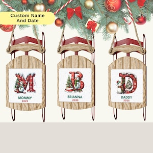 May include: Three personalized Christmas sled ornaments. Each ornament has a wooden sled design with a red frame and a rope for hanging. The center of each sled features a letter with a snowman and Christmas tree design. The text "Mommy 2025", "Brianna 2025", and "Daddy 2025" are displayed.