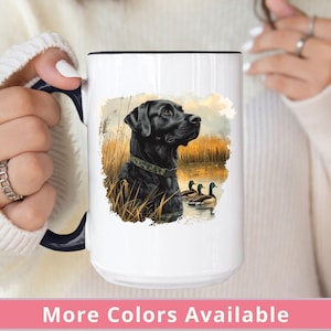 May include: White ceramic mug with a black handle featuring a black Labrador Retriever with a green collar standing in a grassy marsh with three ducks in the water. The mug has a black rim.