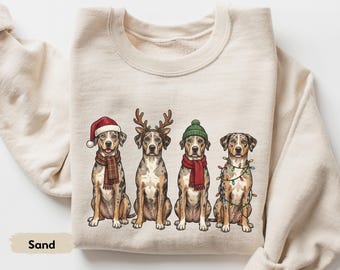 Op de afbeelding: Een zandkleurige sweater met ronde hals en een afbeelding van vier gevlekte honden die op een rij zitten. De honden dragen feestelijke accessoires, waaronder een rode kerstmuts, rendiergeweien, een groene gebreide muts en kerstverlichting.