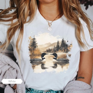 Cozy white colored tshirt featuring a pair of loons on a serene lake with a retro watercolor-style forest and mountain background. Perfect for nature lovers or as a thoughtful vintage-inspired gift.
