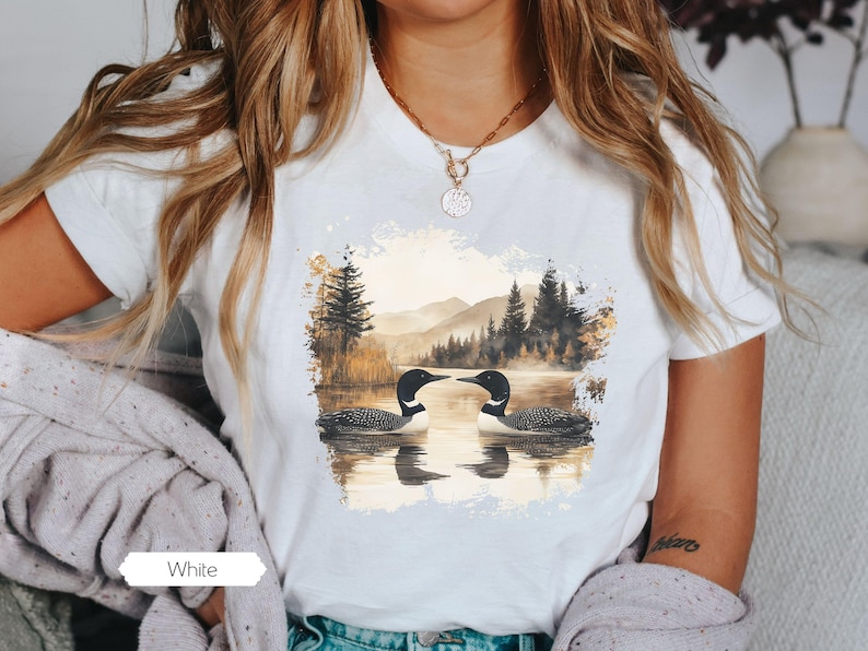 Cozy white colored tshirt featuring a pair of loons on a serene lake with a retro watercolor-style forest and mountain background. Perfect for nature lovers or as a thoughtful vintage-inspired gift.