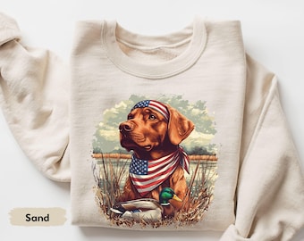 Può includere: Felpa color sabbia con una grafica di un cane marrone con una bandana e una fascia con la bandiera americana. Il cane si trova in un ambiente paludoso con un'anatra. La parola "Sand" è stampata in basso a sinistra.