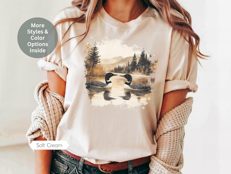 Cozy cream colored tshirt featuring a pair of loons on a serene lake with a retro watercolor-style forest and mountain background. Perfect for nature lovers or as a thoughtful vintage-inspired gift.