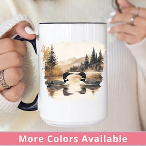 May include: White ceramic coffee mug with a black handle. The mug features a watercolor illustration of two loons facing each other in a lake setting with trees and mountains in the background.