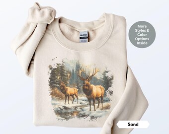 Puede incluir: Una sudadera beige con un estampado gráfico de dos alces en una escena de bosque nevado. El texto "Sand" es visible en la esquina inferior derecha de la imagen.