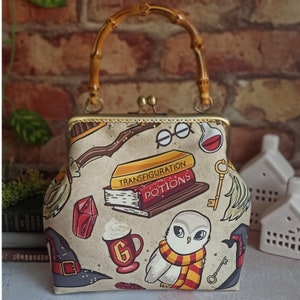 May include: A beige fabric purse with a gold metal frame and a brown wooden handle. The purse features a Harry Potter themed design with illustrations of a broom, a book titled "Transfiguration", a book titled "Potions", a white owl, and a witch's hat.