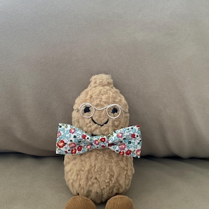 May include: A brown plush toy shaped like a peanut wearing round silver glasses and a bow tie with a floral pattern in red, blue, and white.