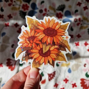 Op de afbeelding: Een sticker met een boeket van drie oranje en rode zonnebloemen met bruine bladeren. De sticker heeft een holografische afwerking.