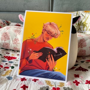 Può includere: Una persona giovane con i capelli biondi sta leggendo un libro nero con una croce bianca sulla copertina. Indossa un maglione rosso e siede su uno sfondo giallo con fiori.